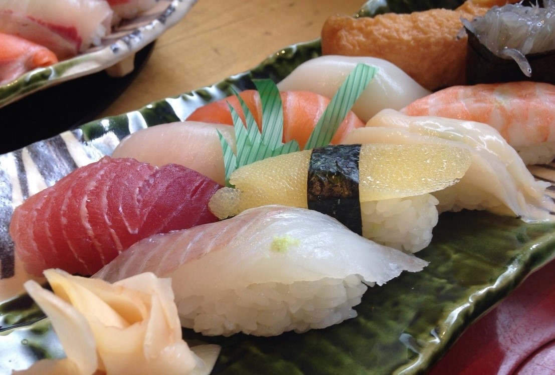 das Bild zeigt – auf einer Schale dekoriert - geformte Bällchen von Sushi-Reis, die lose mit einer Scheibe rohem Fisch oder Meeresfrüchten belegt sind